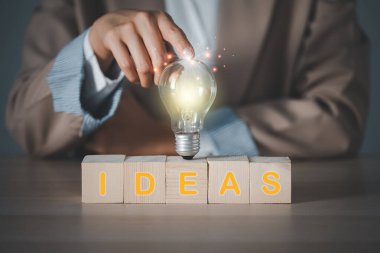 woman's finger touching light bulb on a wooden block with Ideas word, innovative and inspiring new ideas