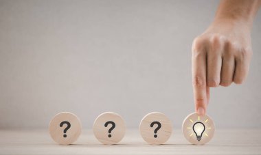 Yakın plan el, ahşap parçayı elinde tutarken ampul simgesi soru işaretleri, düşünce ve fikir kavramları arasında