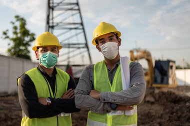 İnşaat alanında çalışan mühendisler. Surat maskesi takan Asyalı işçiler inşaat alanında çapraz kollar takıyor..