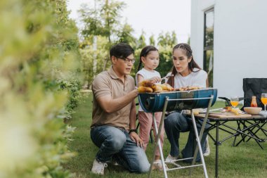 Asyalı bir aile evlerinin arka bahçesinde yemek yiyor. Küçük çocuklu mutlu bir aile kamp yapıyor ve evin arka bahçesinde eğleniyor. Barbekü zamanı, aile aktiviteleri kavramı.