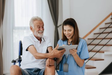 Asyalı hemşire ve kıdemli adam evdeki dijital tablette tıbbi kayıtlara bakıyor. Yaşlı adam ve hemşire sağlık planı için dijital tablet kullanıyorlar. Bakım evi