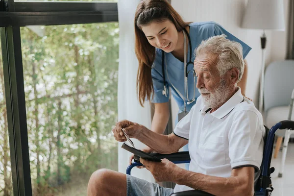 Genç Asyalı hemşire yaşlı adama evde yardım ediyor. Yaşlı bir adam tekerlekli sandalyede oturuyor ve evde genç bir doktorla konuşuyor. Kıdemli sağlık hizmetleri.