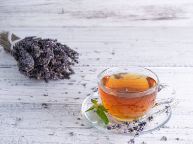Lavantalı çay şeffaf cam bardakta, açık ahşap bir masada. Arka planda bir buket kurutulmuş lavanta.