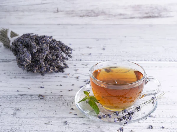 Lavantalı çay şeffaf cam bardakta, açık ahşap bir masada. Arka planda bir buket kurutulmuş lavanta.
