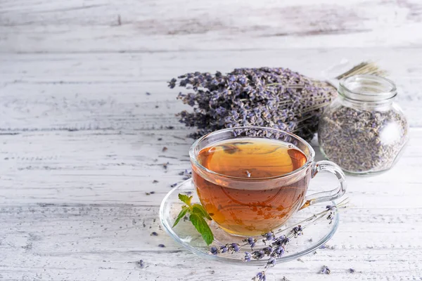 Lavantalı çay şeffaf cam bardakta, açık ahşap bir masada. Bir fincan tabağında bir tutam nane. Arka planda bir kavanoz kurutulmuş lavanta çiçeği.