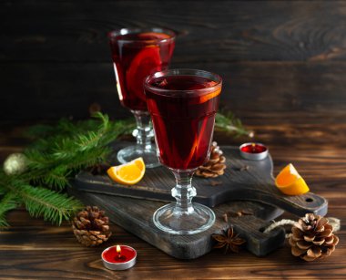 Portakallı sıcak şarap, tarçınlı yıldız anasonlu cam bardaklar ve siyah tahta tahtalar. Arka planda dallar, koniler, mumlar ve mandalina yarıları var.