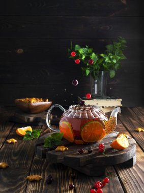 Koyu kahverengi ahşap bir standın üzerinde şeffaf bir çaydanlıkta çilek-narenciye çayı. Ahşap kasede kurabiyeler ve arka planda nane yaprakları. Böğürtlenler çaydanlıklara uçar, etrafa sıçrar