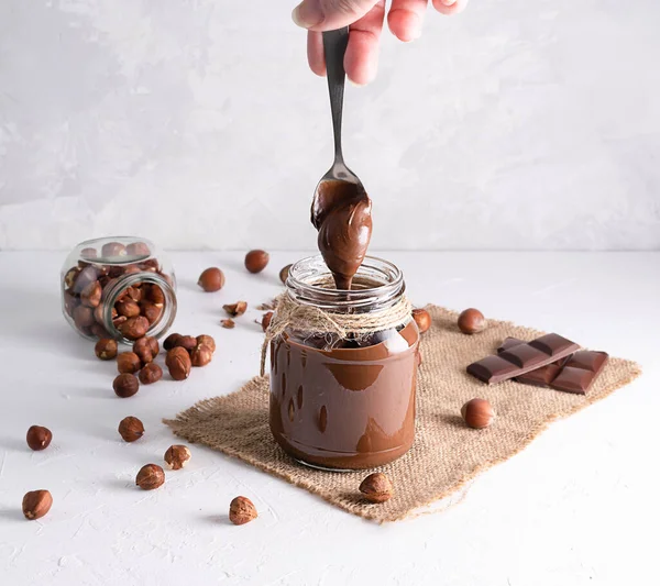 Chocolate-nut paste in a glass jar on a light table. With a spoon, the paste pours into the jar