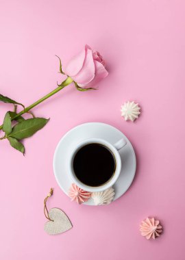 Black aromatic coffee in a white cup on a pink table. White and pink meringue cookies next to the cup. Pink rose in the background. Top view