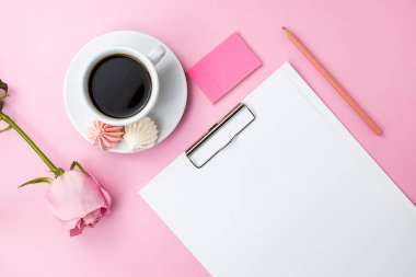 Kağıt, pembe kalem ve açık pembe masa üzerindeki yer imleriyle dolu beyaz bir tablet. Arka planda kahve ve kremalı kurabiye olan beyaz bir fincan. Işık fincanın yanında pembe bir güldür. Üst görünüm