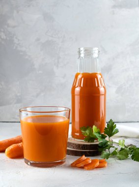 Aydınlık bir masada temiz bir bardakta havuç suyu. Arka plandaki ahşap tezgahta bir şişe meyve suyu. Masada havuç ve maydanoz yaprakları var. Hafif arkaplan