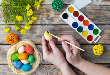 Hasır bir sepette yumurtalar, sarı çiçekler ve ahşap bir arka planda boyalar. Eller yumurtayı sarıya boyar. Paskalya hazırlığı. Üst görünüm