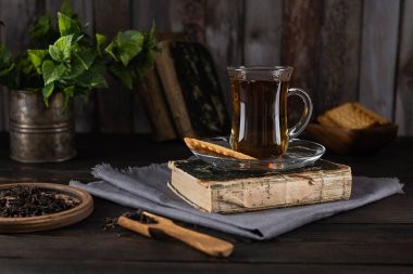 Açık gri peçetedeki eski bir kitaptaki cam bardaktaki çay. Nane yaprakları teneke bardakta ve kurabiyeler arka plandaki tahta kasede. Ahşap arkaplan