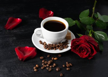Siyah bir masada, beyaz bir fincanda sade kahve. Arka planda kırmızı bir gül ve taç yaprakları. Kahve çekirdekleri masada.
