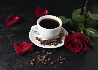 Siyah bir masada, beyaz bir fincanda sade kahve. Arka planda kırmızı bir gül ve taç yaprakları. Kahve çekirdekleri masada.