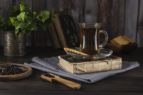 Açık gri peçetedeki eski bir kitaptaki cam bardaktaki çay. Nane yaprakları teneke bardakta ve kurabiyeler arka plandaki tahta kasede. Ahşap arkaplan