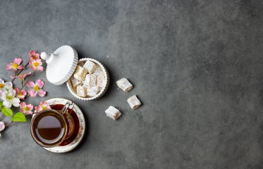 Türk fincanındaki çay ve siyah masadaki beyaz vazoda Türk lokumu. Fincanın yanında bahar çiçekleri var. Üst görünüm, metin için boşluk