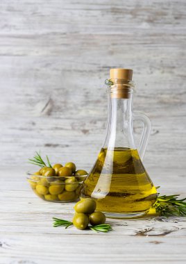 Hafif ahşap bir masada, ertelenmiş bir sürahide altın zeytin yağı. Arka planda cam bir vazoda olgun sulu zeytinler. Hafif ahşap arkaplan