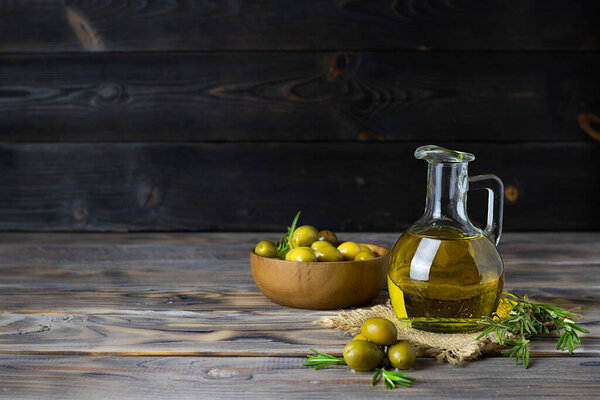 A small glass jug with fresh olive oil on a light canvas napkin. Green olives with a bone in a wooden bowl in the background. Sprigs of rosemary next to the jug