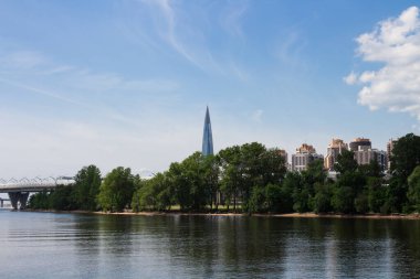 Su manzarası, Lakhta merkezi, köprü, ağaçlar ve mavi arka plandaki konut binaları.