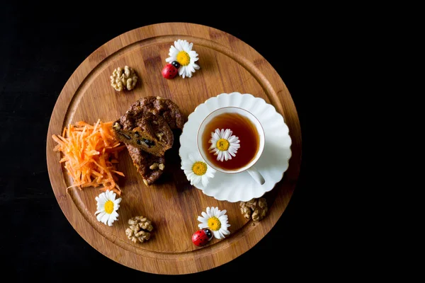 Top view of carrot cakes in a section with walnuts and spices,a white porcelain Cup of tea, walnuts, daisies with ladybirds and grated carrots. All on a round wooden Board on a black background.