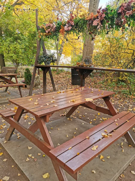 A brown wooden table and benches of an outdoor cafe, covered with fallen leaves, in an autumn park on the Elagin Island of St. Petersburg.