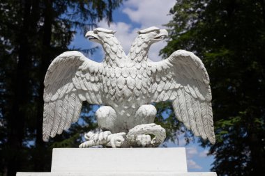 Beyaz çift başlı metal kartal, farklı yönlere bakıyor, ağaçların arka planına ve bulutlu mavi gökyüzüne karşı....