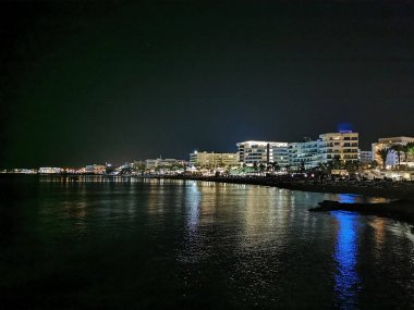 Geceleyin toprak setten Protaras 'a bakın: kumsal, gezinti güvertesi, deniz ve oteller siyah gökyüzünün arka planına karşı.