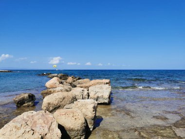 Protaras. Kıbrıs. Akdeniz kıyısından sahile kadar uzanan bir bank ve güneş gölgesi, deniz manzarası, dalgalar ve bulutlu mavi gökyüzü..