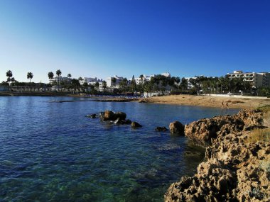 Protaras. Famagusta bölgesi. Kıbrıs. Bulutsuz bir günde Akdeniz körfezinde güneşli şezlonglar ve güneşli, seyrek nüfuslu küçük bir kumsal manzarası..