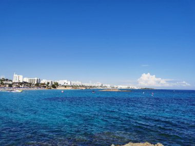 Protaras. Kıbrıs. Akdeniz kıyısında yeşil bitkiler ve taşlar, İncir Ağacı Koyu 'nun kumlu sahili ve bulutlu mavi bir gökyüzünün arka planındaki ada..