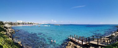 Protaras. Kıbrıs. Akdeniz kıyısında yeşil bitkiler ve taşlar, İncir Ağacı Koyu 'nun kumlu sahili ve bulutlu mavi bir gökyüzünün arka planındaki ada..