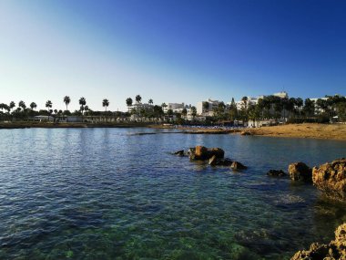 Protaras. Famagusta bölgesi. Kıbrıs. Bulutsuz bir günde Akdeniz körfezinde güneşli şezlonglar ve güneşli, seyrek nüfuslu küçük bir kumsal manzarası..