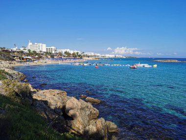 Protaras. Kıbrıs. 10.19.2021 Akdeniz kıyıları yeşil bitkiler ve taşlarla kaplı, İncir Ağacı Koyu 'nun kumlu sahili ve bulutlu mavi bir gökyüzünün arka planındaki ada..