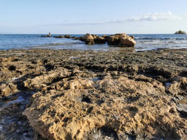 Protaras. Famagusta bölgesi. Kıbrıs. Akdeniz kıyıları, uzun sert lavlar, üzerinde bir balıkçının, mavi gökyüzüne karşı denizde bir eğlence teknesinin bulunduğu taş bir sırt..