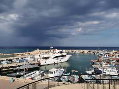 Protaras. Kıbrıs. Rıhtım, Akdeniz körfezinde küçük gemileri, yatları ve tekneleri olan liman dramatik bir gökyüzünün arka planına karşı..