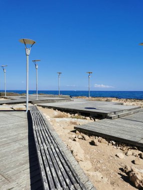 Protaras. Famagusta bölgesi. Kıbrıs. Akdeniz 'in kayalık kıyıları boyunca tahta bir bankı ve fenerleri olan bir tahta kaldırım..