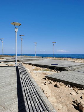 Protaras. Famagusta bölgesi. Kıbrıs. Akdeniz 'in kayalık kıyıları boyunca tahta bir bankı ve fenerleri olan bir tahta kaldırım..