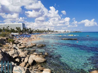 Protaras. Famagusta bölgesi. Kıbrıs. Kıyıya yakın büyük taşlar, Akdeniz 'in berrak suları, kumlu büyük Sunrise plajı manzarası ve bulutlu mavi gökyüzünün arka planında oteller..