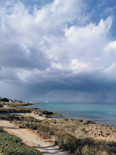 Protaras. Famagusta area. Cyprus. Sunny coast of the Mediterranean Sea with grass and stones, a dramatic sky and rain over the sea, a boat in the sea and the largest sailing yacht in the world.