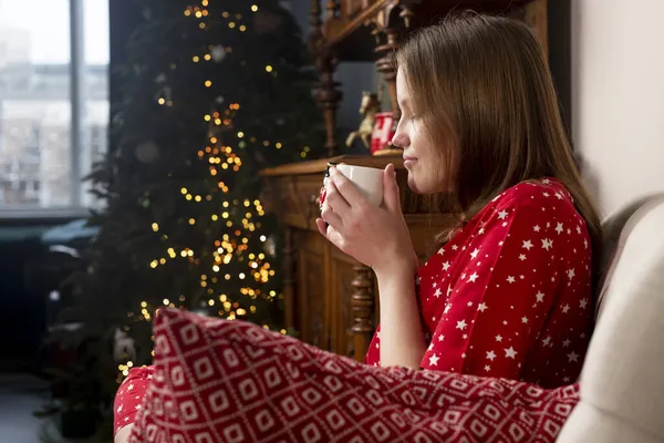 Mujer con taza de té en las manos, sentado en silla en casa acogedora con abeto en las vacaciones de Navidad —  Fotos de Stock