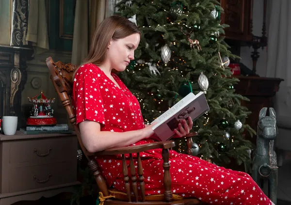 Mujer relajante con libro en mecedora con árbol de Navidad en el fondo en las vacaciones de invierno. Lectura de la persona en casa acogedora por la noche —  Fotos de Stock