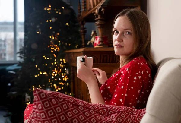 Retrato de mujer joven con taza de té caliente sentado en la silla en casa hygge Scandi en las vacaciones de invierno con abeto y luces —  Fotos de Stock