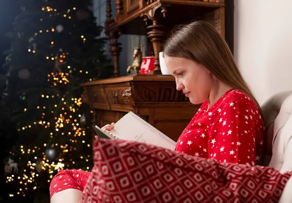 Mujer leyendo libro, sentado en la silla cerca del árbol de Navidad en la víspera de Navidad en casa acogedora —  Fotos de Stock