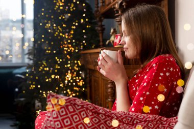 Elinde çay fincanı olan bir kadın, Noel tatilinde köknar ağacıyla rahat bir evde sandalyede oturuyor.