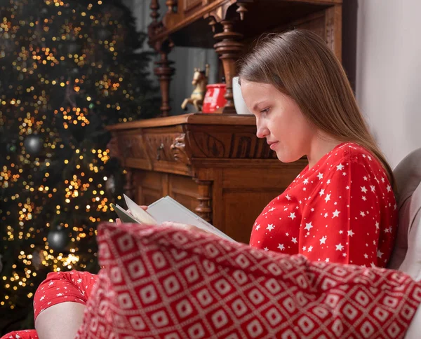 Mujer en ropa de casa roja leyendo libro de Navidad en silla con abeto en las vacaciones de Navidad —  Fotos de Stock