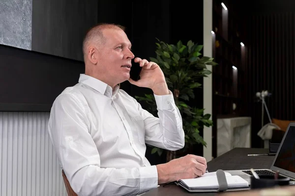 Geschäftsanruf im Büro. Geschäftsmann sitzt mit Laptop am Schreibtisch, telefoniert und macht sich Notizen im Planer. — Stockfoto