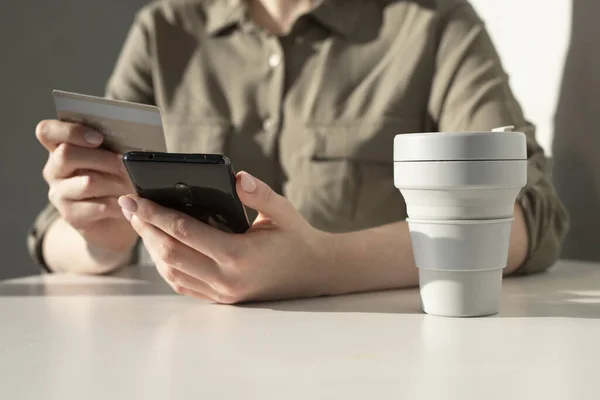 Mãos femininas segurando cartão de crédito e telefone. Pagamento on-line no conceito de aplicativo. Mulher sentada à mesa com taça takeaway e compras online Fotografia De Stock