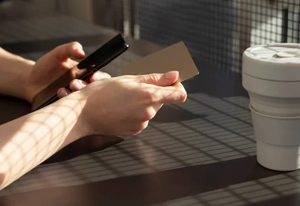 Mãos femininas segurando cartão de crédito e smartphone. Pagamento online ou conceito de compras. Mulher sentada na mesa no café com caneca e informações de entrada de celular — Fotografia de Stock