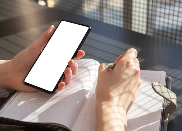 Femme mains tenant téléphone maquette et stylo et prendre des notes dans le planificateur. Femme assise au bureau et écrivant des informations ou un ordre du jour à partir de l'écran du smartphone — Photo